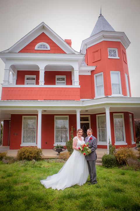 Bride in flowing white wedding dress with her husband in front of red house.